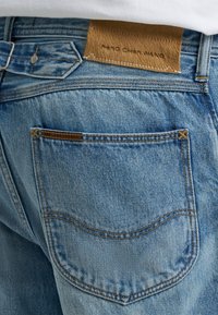 Ανοιχτό μπλε τζιν από denim με πίσω τσέπη και δερμάτινη ετικέτα επωνυμίας «PENG CHEN WANG» ραμμένη πάνω από τη μέση και στην άκρη της τσέπης.