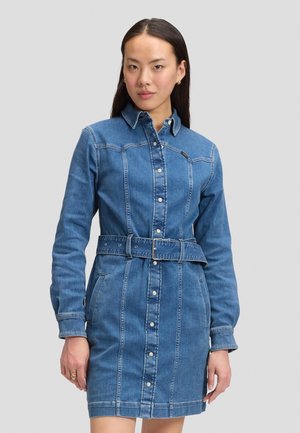 Donna che indossa un vestito di jeans blu a maniche lunghe con chiusura a bottoni e cintura abbinata, in piedi davanti a uno sfondo grigio chiaro uniforme.