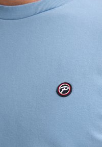 T-shirt in cotone azzurro chiaro con un'etichetta rotonda e testurizzata in nero e rosso sul lato sinistro del petto. Texture liscia, scollo a girocollo.