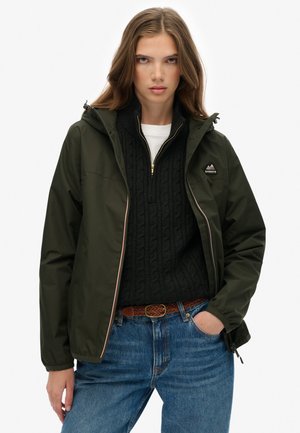 Veste zippée vert olive avec capuche, portant un pull en laine noir en dessous. La veste a un logo sur la poitrine et des poignets élastiques.