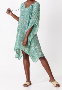 Kaftan šaty v zeleném paisley vzoru, vyrobené z lehké tkaniny. Mají výstřih do V a volné, splývavé rukávy. Kombinované s plochými sandály.
