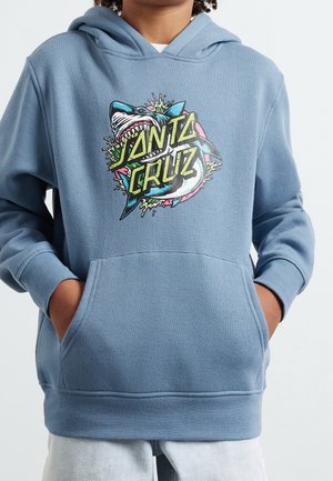 Młoda osoba w niebieskiej bluzie z kapturem z rękami w przedniej kieszeni, z nadrukiem rekina i napisem "Santa Cruz" na piersi.