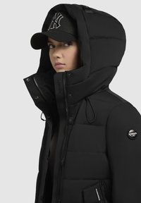 Crna puffer jakna s visokim ovratnikom i kapuljačom s vezicom, koja ima patentni zatvarač sprijeda, bočne džepove i oznaku s logotipom na rukavu.