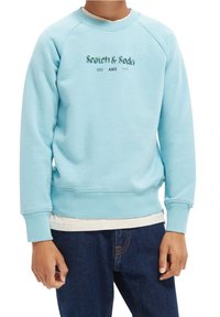 Sudadera azul claro con mangas raglán, puños acanalados y cuello redondo. Presenta "Scotch & Soda AMS" impreso en el frente.