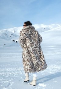 Leopárdmintás műszőrme kabát széles gallérral, egy hóval borított tájban látható, fehér nadrággal és világos csizmával párosítva.