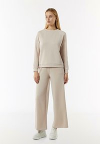 Beige bijpassende sweatshirt en wijde broek in zachte stof. De sweatshirt heeft een ronde halslijn en geribde manchetten. Schoenen zijn witte sneakers.