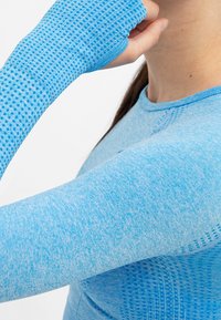 Närbild på en person som har på sig en blå strukturerad långärmad sporttröja med tumhål, som rör vid sin nacke.