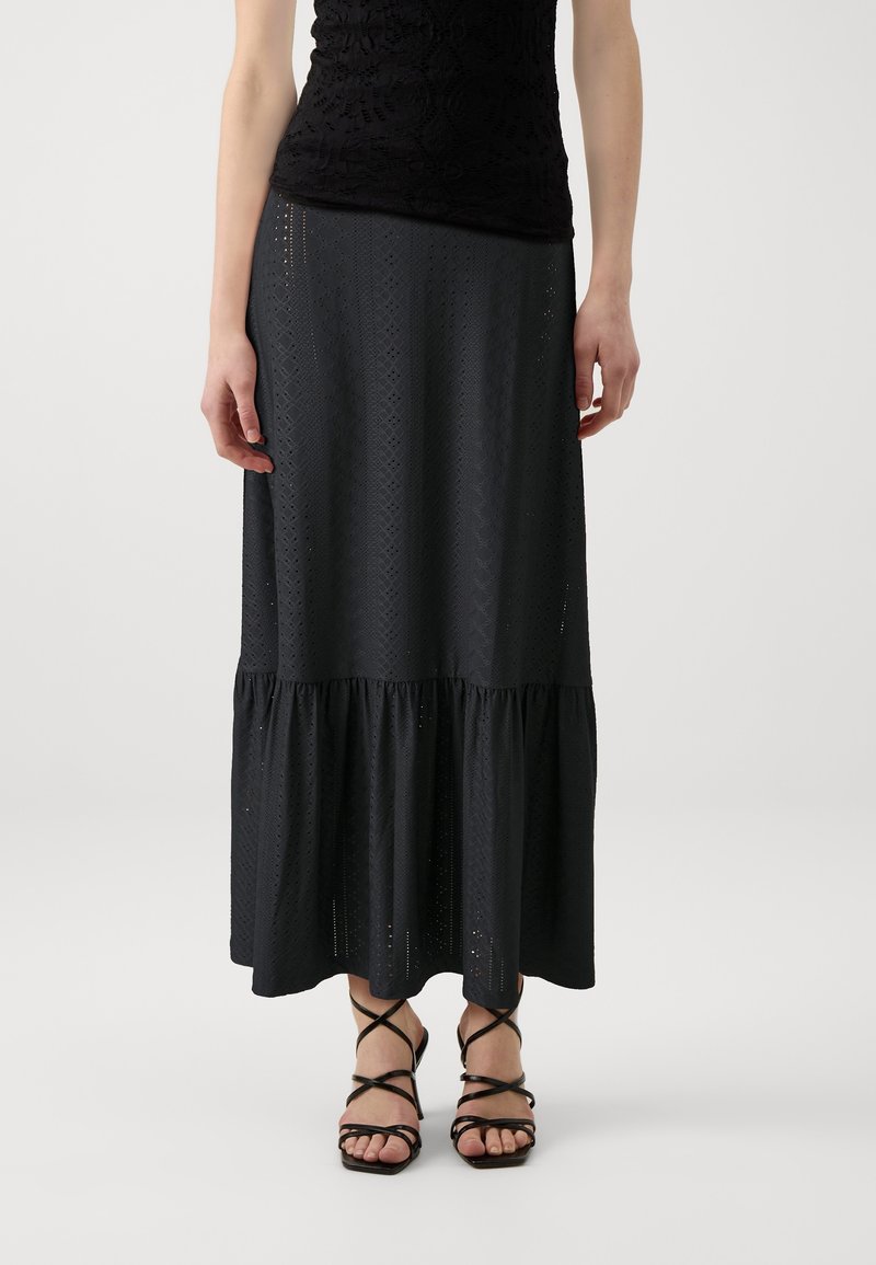Jupe noire en œillets avec un ourlet à volants, présentant un motif texturé et un tissu léger. Portée avec des sandales noires à lanières.