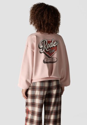 Woman with curly hair wearing a pink sweatshirt with "Rodeo live Las Vegas" text and plaid pants facing away against a plain background.