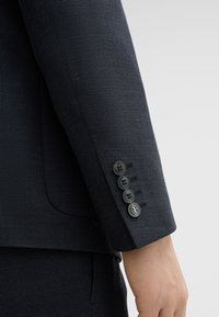 Manica di giacca scura su misura con un tessuto texture. Presenta quattro bottoni neri e un design minimalista. Nessun motivo o accento visibile.