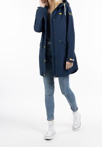 Trench-coat imperméable bleu marine avec capuche, accents jaunes, deux poches avant et fermeture éclair, associé à un jean bleu et des baskets blanches.