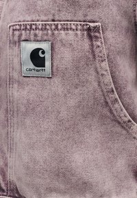 Κοντινό πλάνο ξεθωριασμένης μωβ τζιν τσέπης με την ετικέτα της μάρκας Carhartt ραμμένη στην επάνω αριστερή γωνία.