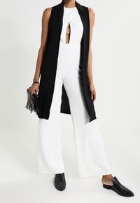 Schwarze ärmellose Weste über einem weißen Jumpsuit mit einem Schlüsselloch-Ausschnitt. Schwarze Mules und eine kleine schwarze Handtasche vervollständigen den Look.
