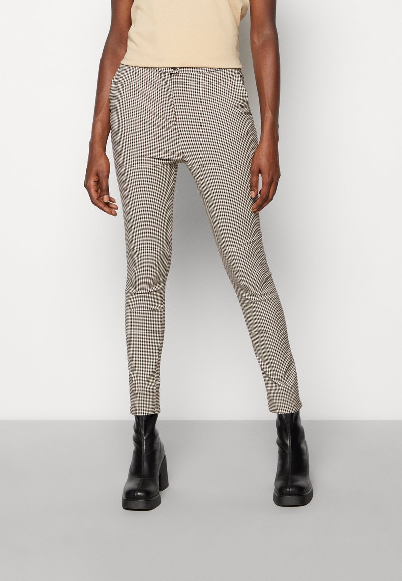 Personne portant un pantalon slim à carreaux beige et noir avec des bottines à talons noires, debout devant un fond clair uni.