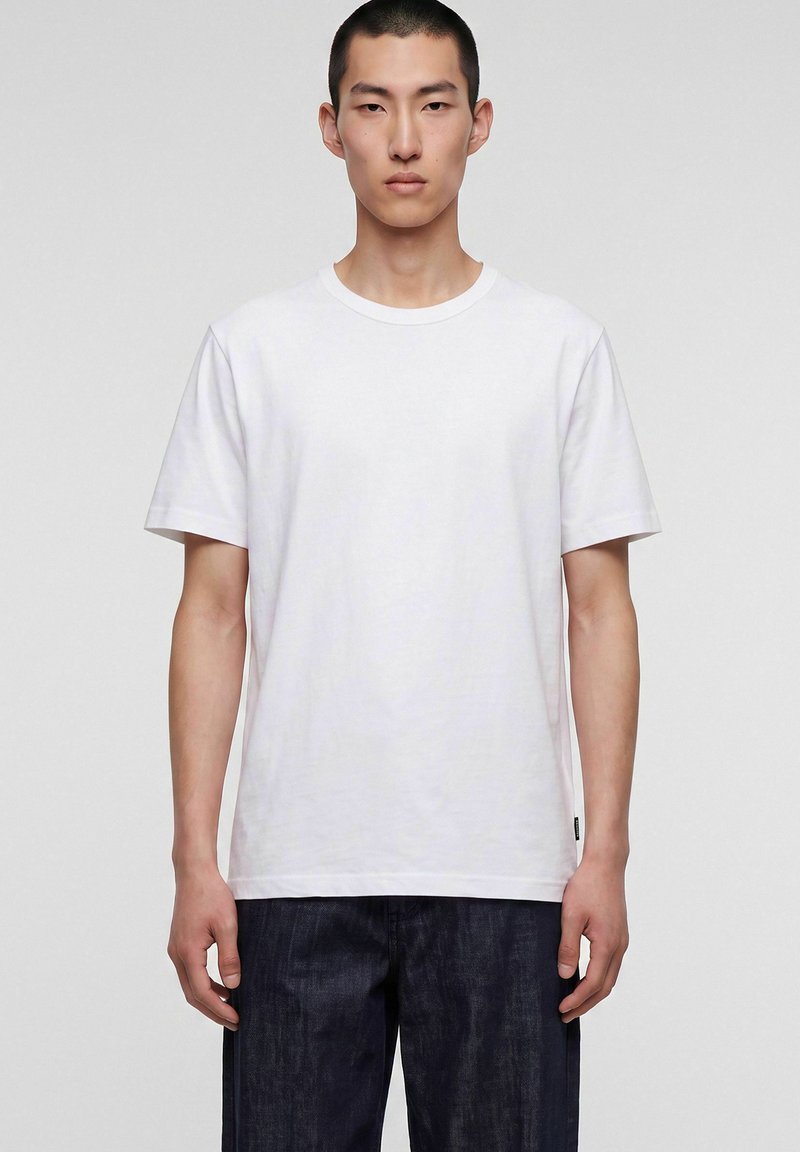 Jeune homme aux cheveux courts portant un t-shirt blanc uni à manches courtes et un pantalon foncé, debout devant un fond clair uni.