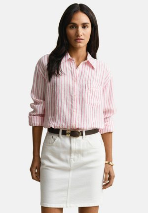 Vrouw draagt een roze-wit gestreept overhemd met knopen, ingestopt in een witte rok met een zwarte riem, staand tegen een effen achtergrond.
