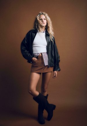 Schwarze Lederjacke in Übergröße, weißes enges Top, gestreifte Bluse, brauter Wildleder-Minirock und knielange schwarze Stiefel vor einem braunen Hintergrund.
