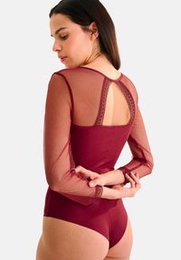 Body en bordeaux avec des manches longues en mesh transparent, un accent en dentelle au dos et une coupe ajustée, soulignant la douceur et l'élégance.