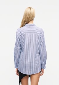 Ξανθιά γυναίκα που φοράει μπλε και λευκό κάθετο ριγέ πουκάμισο με μακριά μανίκια και μαύρη ασύμμετρη φούστα, στέκεται πλάτη στην κάμερα.