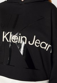 Czarna bluza z kapturem, z błyszczącym czarnym napisem "Klein Jeans" i odważnym wzornictwem. Miękka tkanina o kroju cropped.