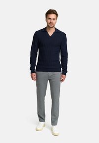 Navy-blaues Rippstrick-Pullover mit V-Ausschnitt und langen Ärmeln, kombiniert mit grauen Hosen und weißen Sneakers. Glatte Textur über das gesamte Kleidungsstück.