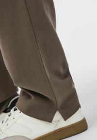 Braune, strukturierte Hose mit maßgeschneidertem Schnitt und seitlichen Schlitzen, kombiniert mit weißen Sneakers, die ein glattes Finish und ein zeitgenössisches Design präsentieren.