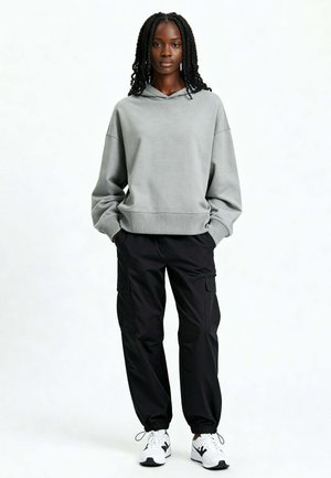 Vrouw met gevlochten haar, gekleed in een loszittende grijze hoodie, zwarte cargobroek en witte sneakers, staand met handen in haar zakken.