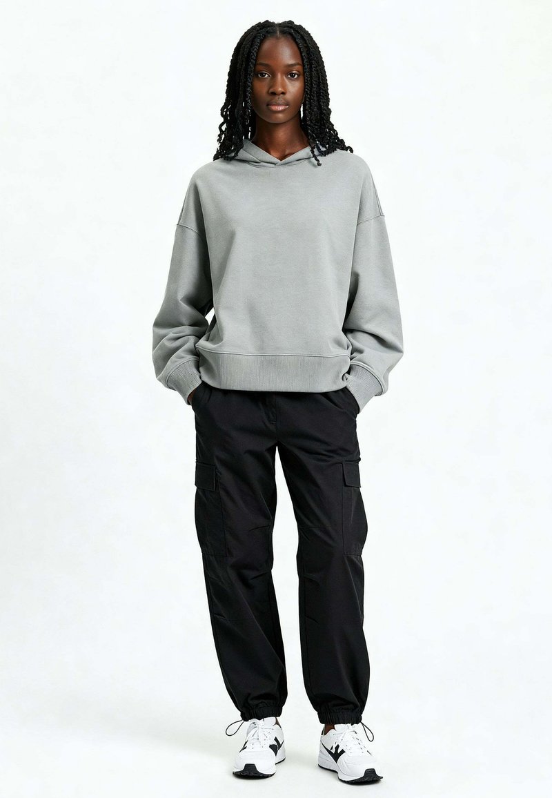 Femme aux cheveux tressés portant un sweat à capuche gris ample, un pantalon cargo noir et des baskets blanches, debout les mains dans les poches.