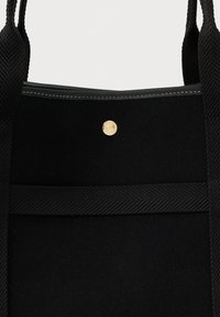 Μαύρη τσάντα ώμου από ύφασμα με υφασμάτινες λαβές, οριζόντια λεπτομέρεια ζώνης, κλείσιμο με κουμπί πρέσας και λευκή ραφή κατά μήκος της πάνω άκρης.