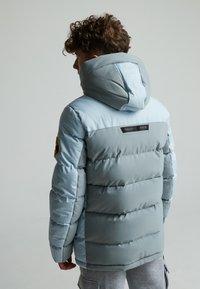 Veste matelassée bleu clair avec capuche, présentant une texture matelassée, un patch logo sur la manche, et une section supérieure de couleur bleu foncé contrastante.