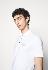 Camisa polo blanca de algodón, con un cierre de tres botones, mangas cortas y un logo gris "EA7" en el pecho.