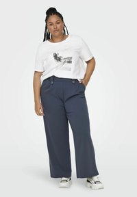 Valkoinen t-paita, jossa on valokuvaprintti, yhdistettynä tummansinisiin leveälahkeisiin housuihin, joissa on etunapit ja sileä pinta. Tenistenteet täydentävät asun.