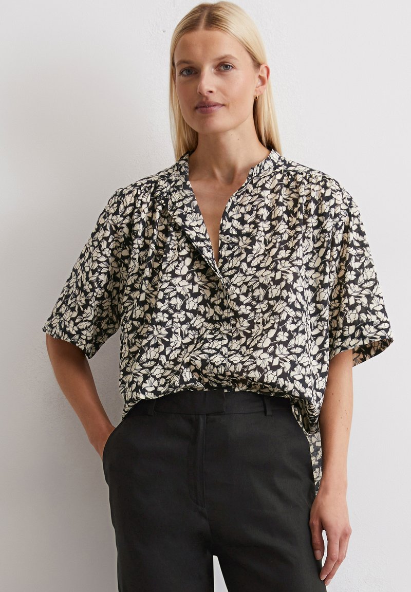 Chemise noire à manches courtes avec un motif floral blanc et beige, dotée d'un col et d'une coupe décontractée. Portée avec un pantalon noir ajusté.