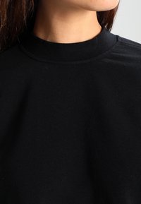 Primer plano del cuello y el hombro que llevan un top de punto negro de cuello alto, contra un fondo claro y liso.