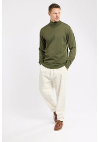 Homme debout les mains dans les poches, portant un pull à col roulé olive, un pantalon blanc et des bottes en cuir marron, regardant sur le côté.