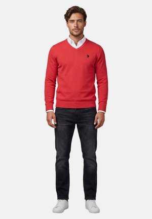 Mann steht frontal, trägt roten V-Ausschnitt-Pullover über weißem Hemd mit Kragen, schwarze Jeans und weiße Turnschuhe vor weißem Hintergrund.