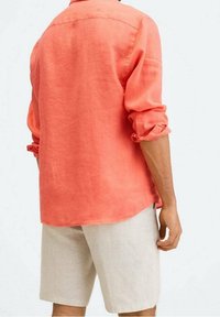 Camicia in lino corallo brillante con maniche arrotolate, dotata di colletto a punte e vestibilità rilassata. Abbinata a pantaloni corti beige, mostrata da dietro.