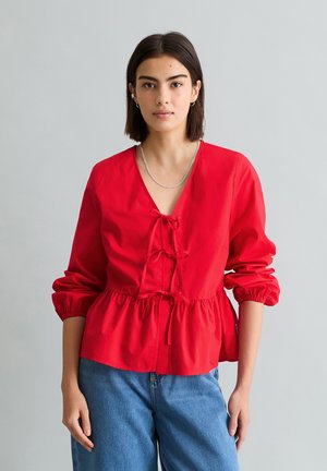 Femme aux cheveux raides et foncés portant un chemisier rouge à manches longues avec des liens devant et un jean bleu, debout devant un fond gris uni.