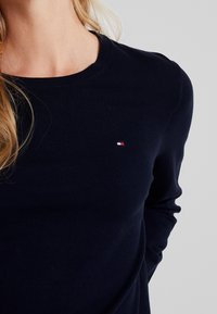 Tmavě modrý pletený svetr s kulatým výstřihem, s malým červeným, bílým a modrým logem na hrudi. Materiál vypadá hladký a přiléhavý.