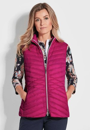 Fuchsia gesteppte Weste mit Reißverschluss vorne, hohem Kragen und Seitentaschen, über einem blumendruckten Hemd mit Dreiviertelärmeln getragen.