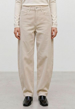 Personne portant un pantalon en velours beige, des mocassins noirs brillants et un haut à manches longues crème, debout sur un sol clair.
