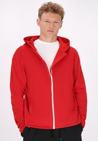 Rode zip-up hoodie met een capuchon, gemaakt van een glanzende stof. Heeft een witte rits en zakken. Draagt over een wit shirt, met een getailleerd ontwerp.