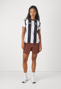 Schwarz-weiß gestreiftes Kurzarm-Jersey mit Logo auf der Brust, kombiniert mit braunen Shorts, die ein Frontreißverschlussdetail aufweisen, und weißen Sneakers.