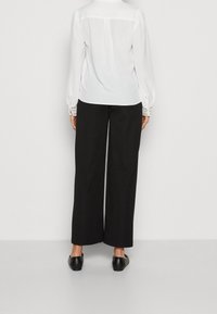 Blouse blanche à manches bouffantes, accents en dentelle aux poignets, associée à un pantalon noir taille haute à jambes larges et à des chaussures noires à enfiler.