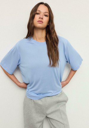 Kvinde med langt brunt hår iført en løs lyseblå T-shirt og grå bukser, stående med hænderne på hoften mod en ensfarvet baggrund.