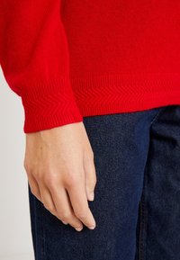 Gros plan d'une main posée à côté d'un jean bleu foncé, portant un pull en tricot rouge avec des poignets et un ourlet texturés en zigzag.