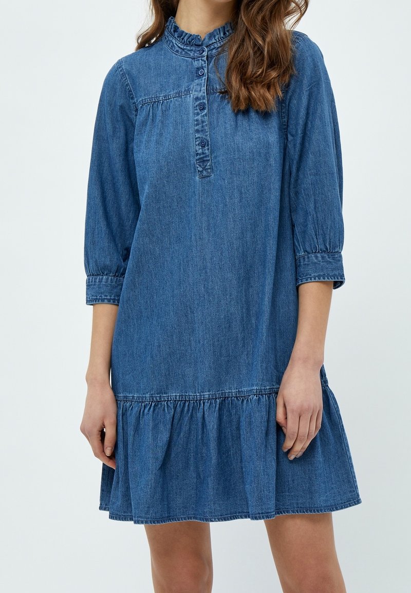 Denim dress with a ruffled neckline, button placket, and three-quarter sleeves. Features a gathered, tiered skirt in medium blue.