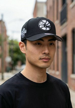 Young man wearing a black 76ers baseball cap and black shirt, standing outdoors in an urban setting with blurred buildings in the background.