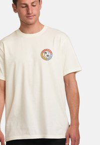 Cream-colored cotton t-shirt featuring a circular graphic logo on the left chest with a multicolored design and text "BILLABONG SINCE 1973."