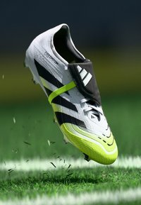 Cizme de fotbal Adidas Predator albe și verzi neon aruncând iarbă pe un teren verde, cu un fundal întunecat.